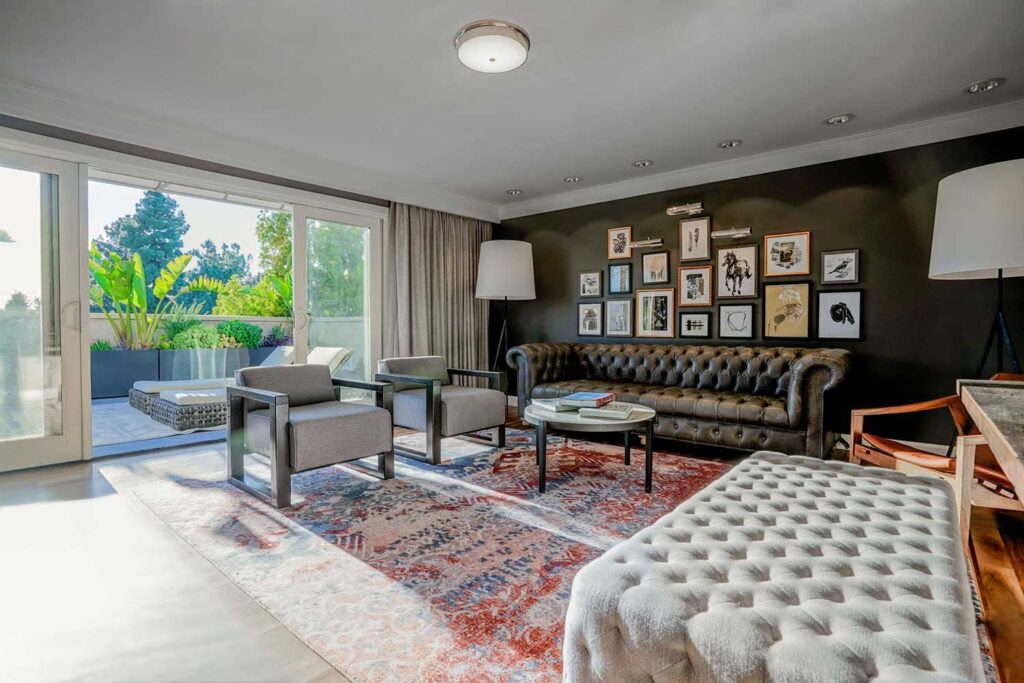 Modern suite living room at Hotel Amarano featuring a tufted leather sofa, two gray armchairs, a patterned area rug, gallery wall art, and large sliding doors opening to a private terrace with lush greenery.