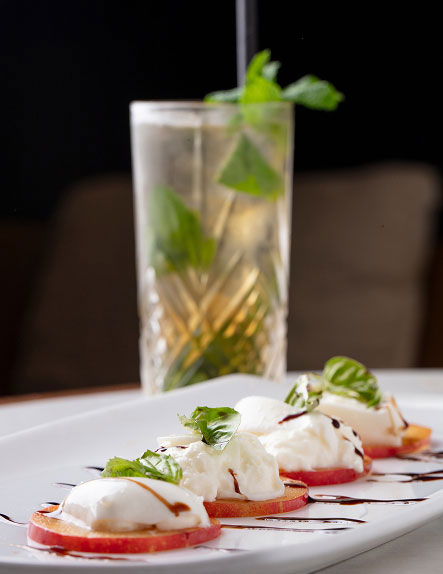 Caprese-style appetizer with fresh mozzarella, sliced tomatoes, basil, and balsamic glaze, served on a white plate with a mint-garnished drink in the background.
